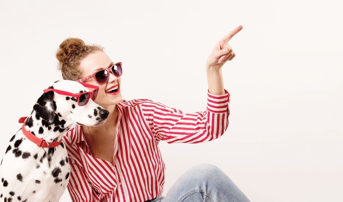 Happy girl and pet in red sunglasses. Young woman hugs dalmatian dog and points on right, isolated on white background. Check this out. Place for text